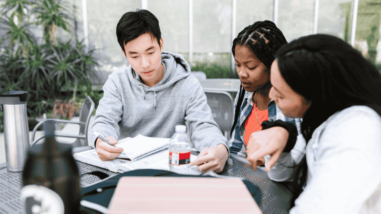 Trois jeunes adultes étudient ensemble à une table, concentrés sur un livre qu’ils lisent en collaboration.