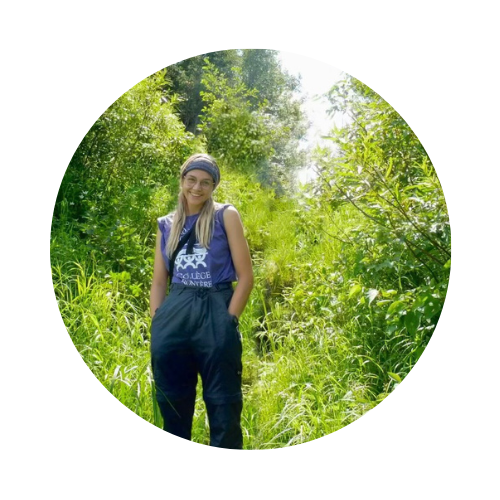 A young woman with blond hair stands in a wooded area.